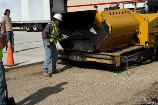 a man in a hard hat and safety vest loading asphalt into a machine